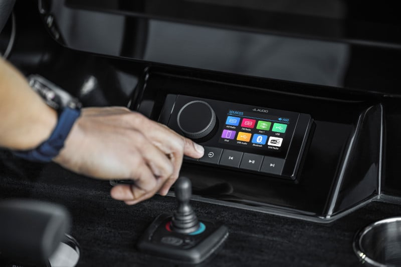 Un bărbat stă în partea din față a bărcii sale de pescuit la bass, controlând motorul electric. Barca sa se află în apropierea țărmului, în timp ce soarele apune în fundal.